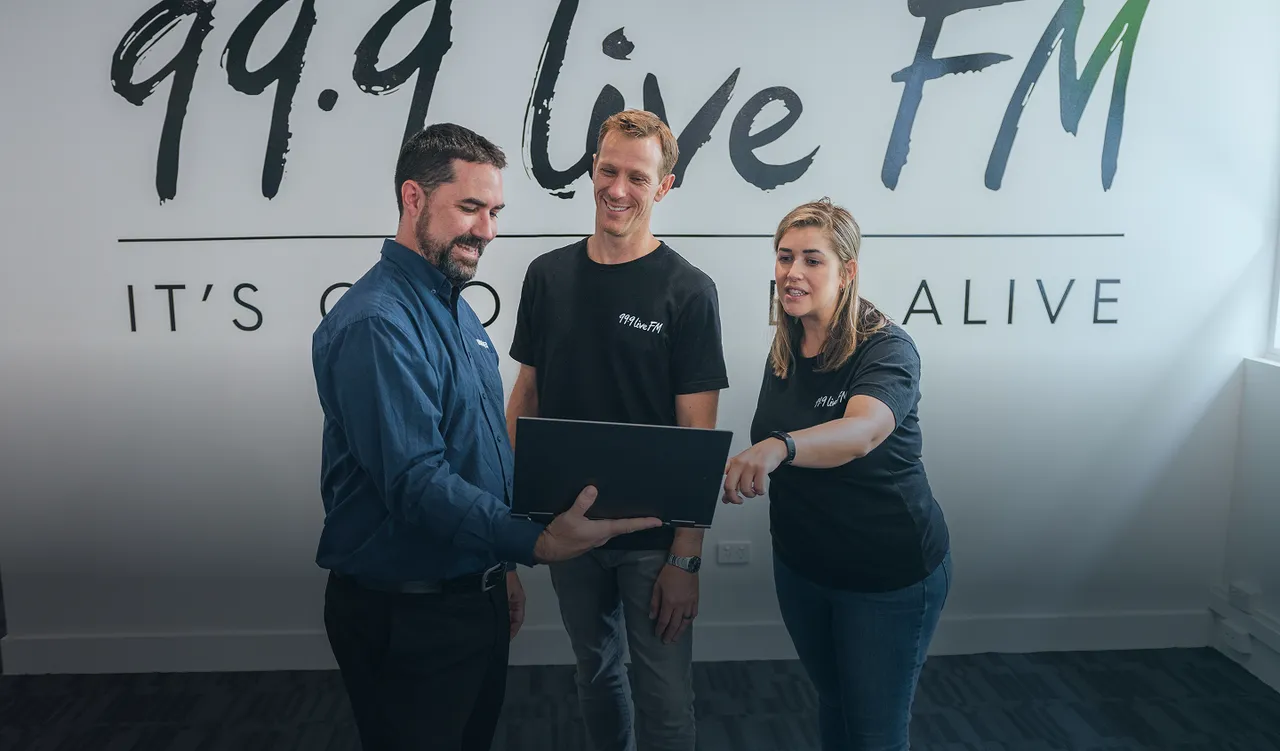 Three DakTech staff looking at a laptop in front of the 99.9 Live FM studio wall